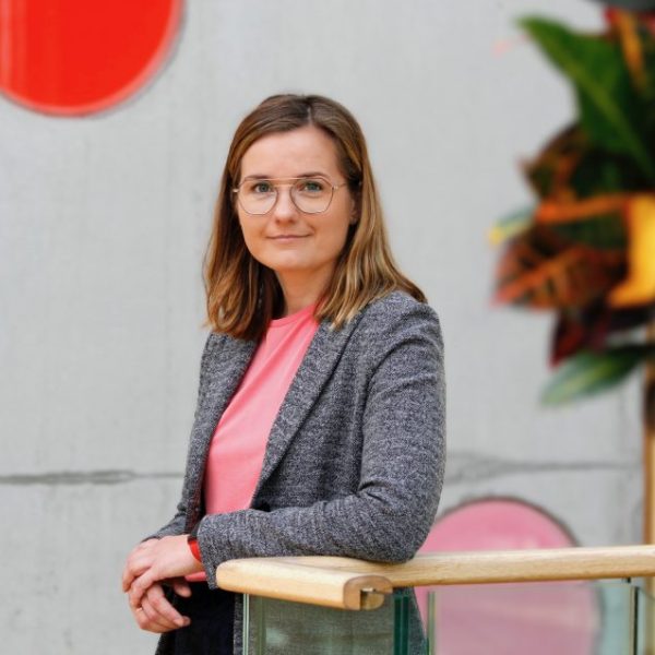 Zanda Martens lächelt in die Kamera. Sie trägt eine Brille mit schwarzem Rahmen, einen grauen Blazer und ein rosa Oberteil. hinter ihr ist eine graue Betonwand zu sehen.