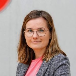 Zanda Martens lächelt in die Kamera. Sie trägt eine Brille mit schwarzem Rahmen, einen grauen Blazer und ein rosa Oberteil. hinter ihr ist eine graue Betonwand zu sehen.
